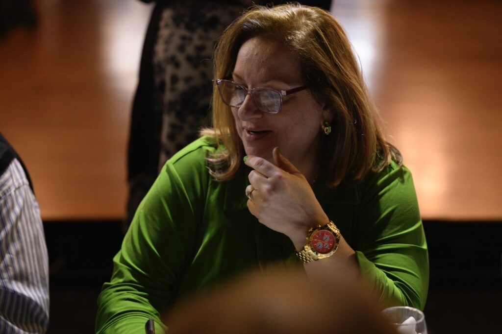A woman in green sits at a table with her hand on her chin.