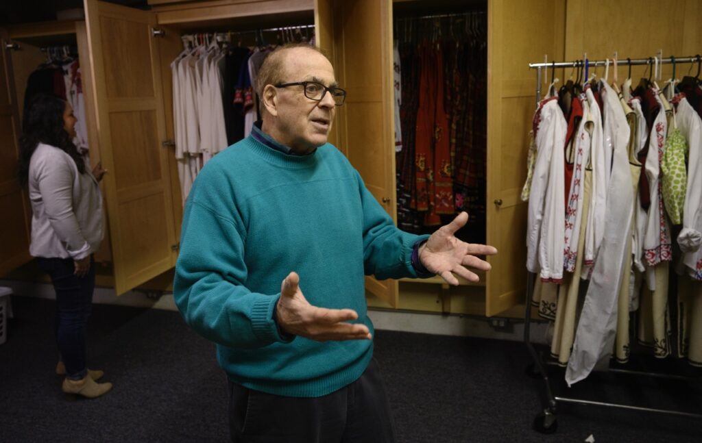 A man in a teal sweater holds out his hands as he speaks.