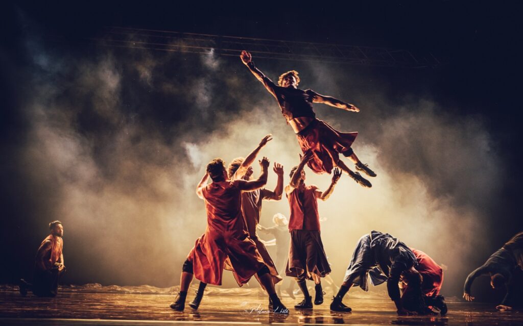 A group of dancers in red hold their hands up to catch another dancer flying through the air.
