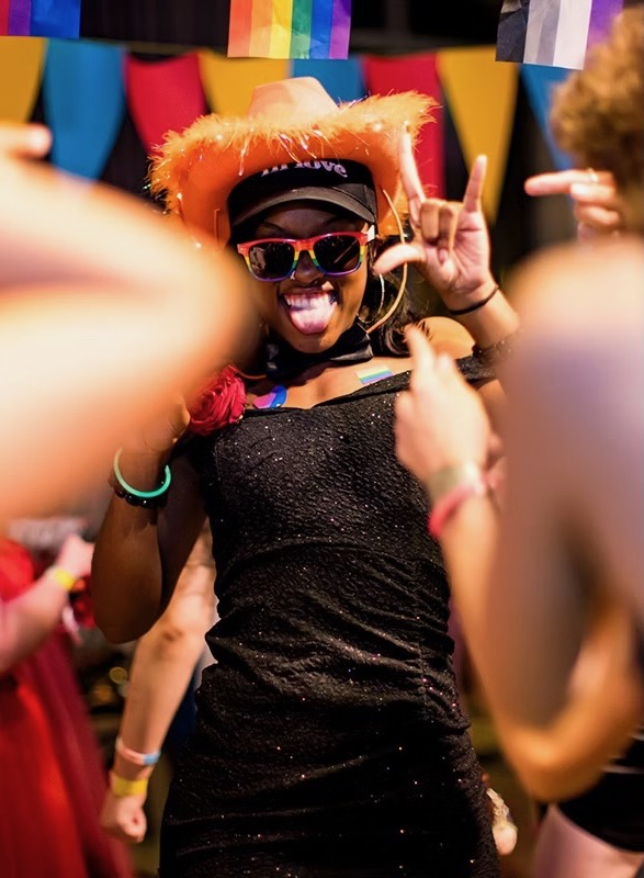 A woman in pride gear and an orange cowboy hat dances with sunglasses on.