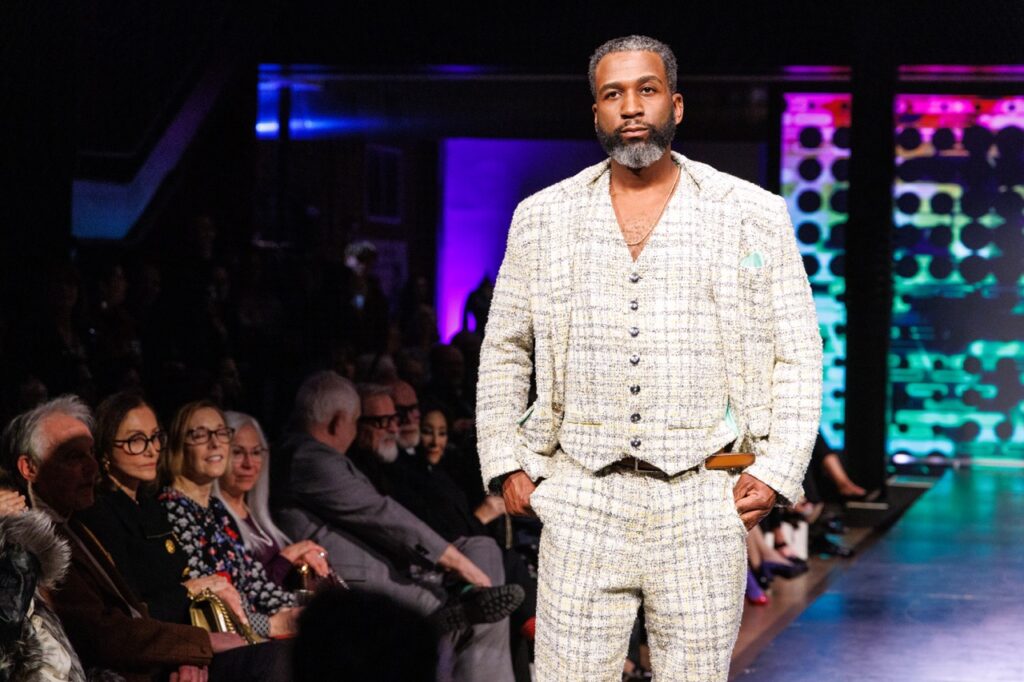 A man in a white suit walks down a fashion show runway.