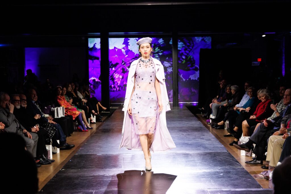 A woman in a pink gown and hat walks down a fashion runway.