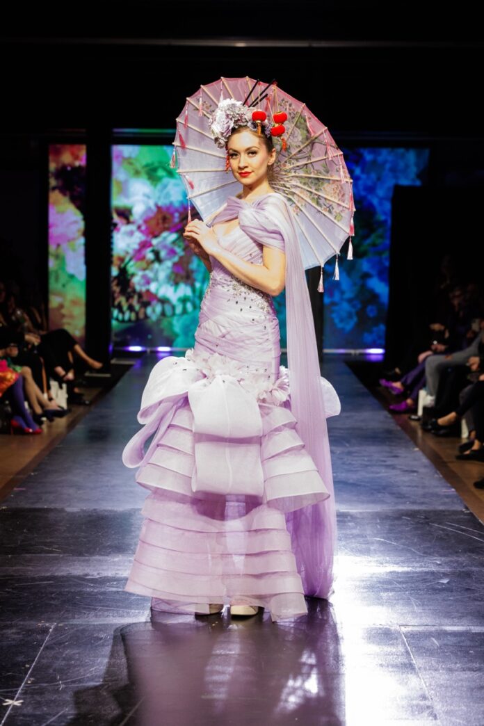 A woman in a lavender gown and parasol on a fashion show runway.