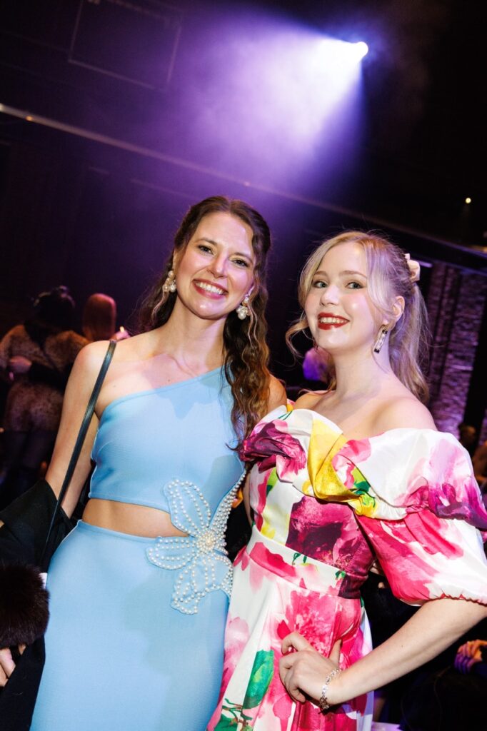 Two women, one in a blue gown and the other in a pink and white gown stand together in a club area.