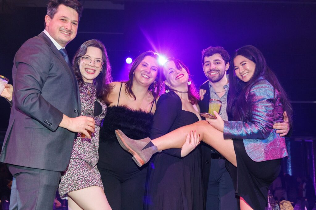 A group of people gather together with drinks in hand under a purple club light.
