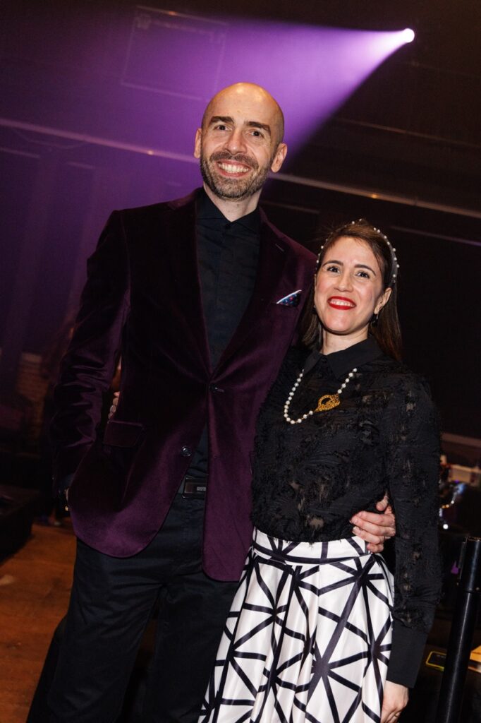 A man in a purple velvet jacket and a woman in a black long sleeve shirt and pattern skirt stand together.