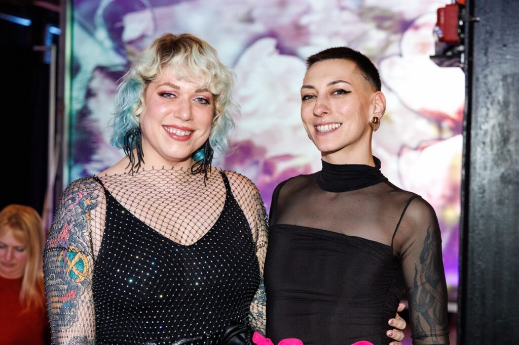 Two women stand arm in arm in front of a watercolor background.