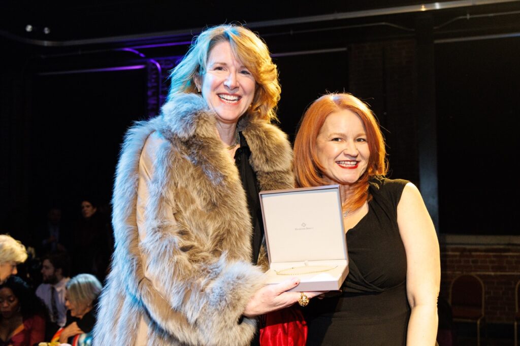 One woman in a fur jacket gives another woman a diamond necklace.