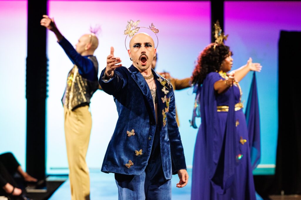 Three people in blue fashion outfits perform on a runway with their hands held out.