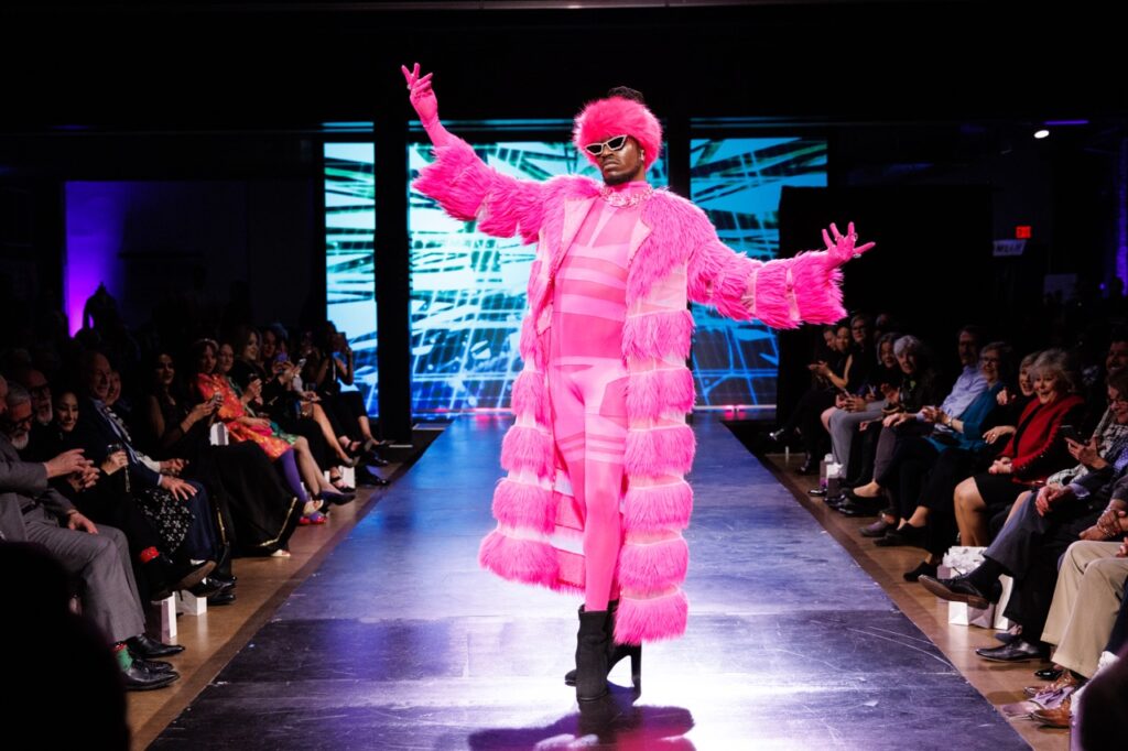 A man in a hot pink bodysuit and fur coat and hat poses at the end of a runway.