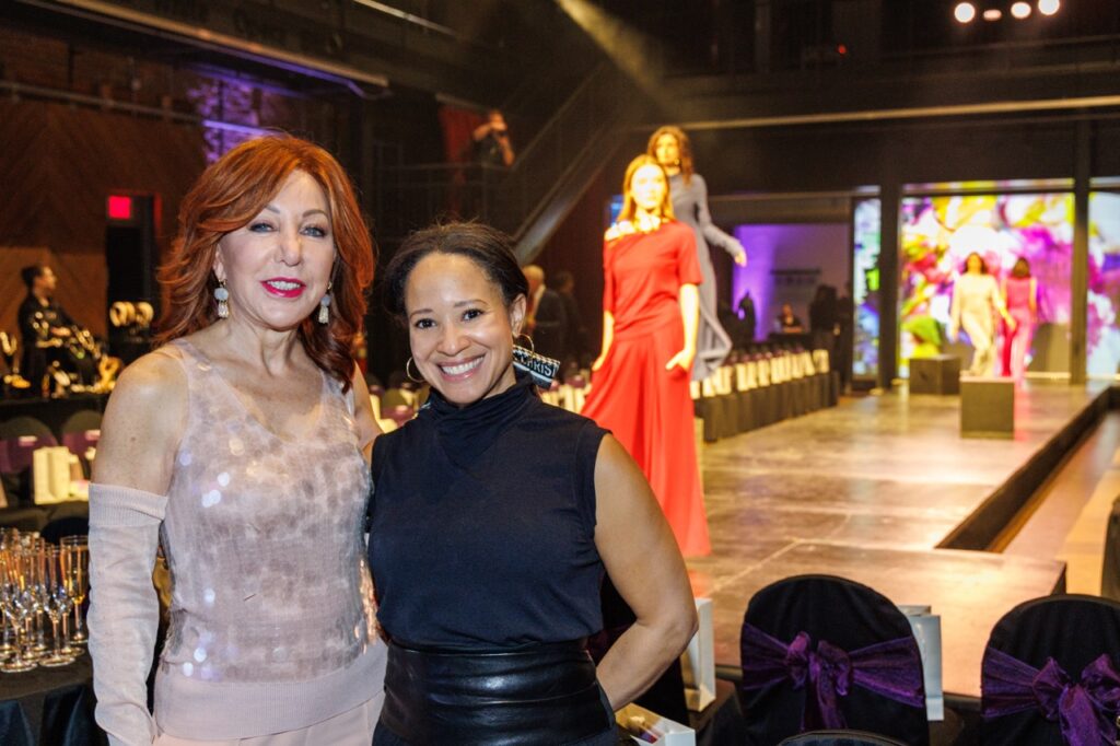 Two women stand arm in arm in front of a fashion show runway with models walking down it.