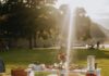 Pittsburgh Businesses to Help Create Your Perfect Picnic A white blanket with pillows lays at a picnic in a Pittsburgh Park with a table over top of it full of picnic lunch foods and flowers.