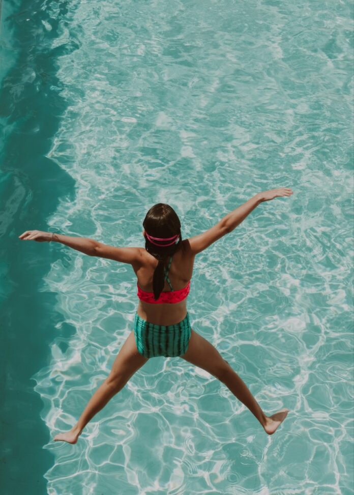 A girl jumps into a swimming pool with a pink and green bathing suit on.