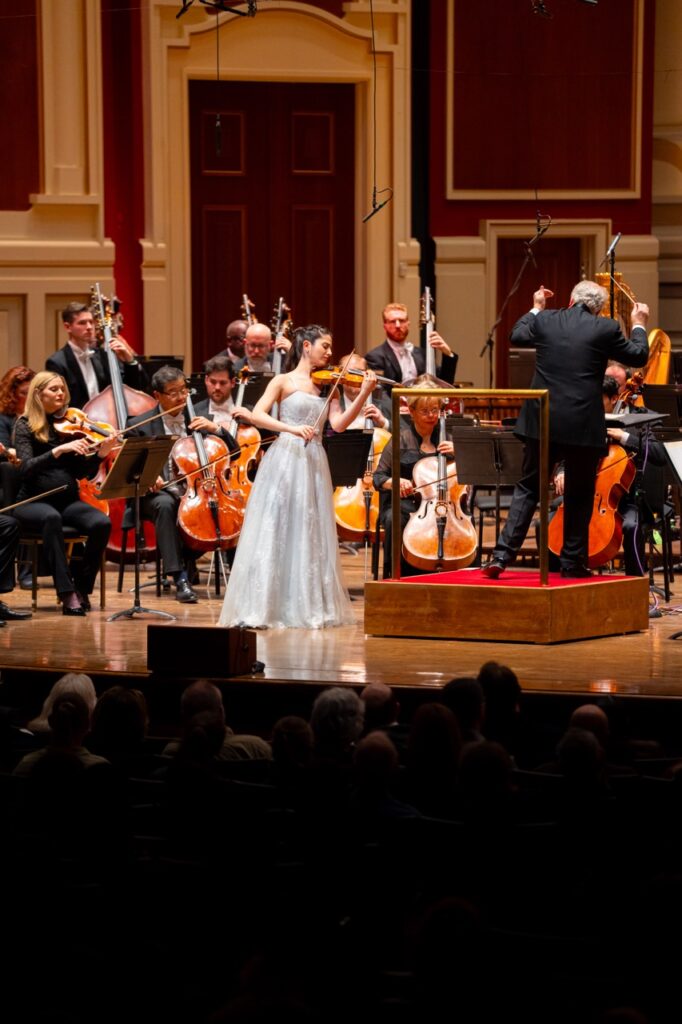 A woman in a long silver dress stands and plays violin in front of a symphony on a stage.