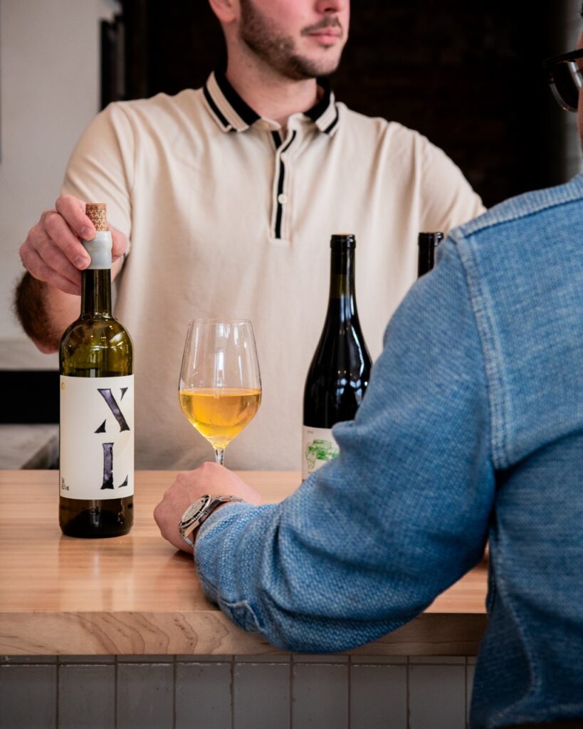 A man pushes forward a bottle and glass of wine across a wine bar.