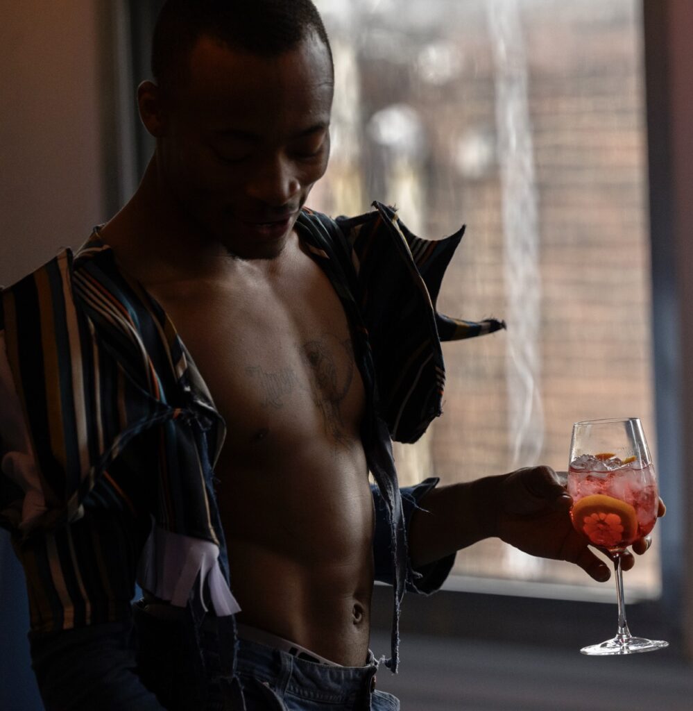 A man with an open jacket and no shirt underneath holds a wine glass with a red cocktail in it.