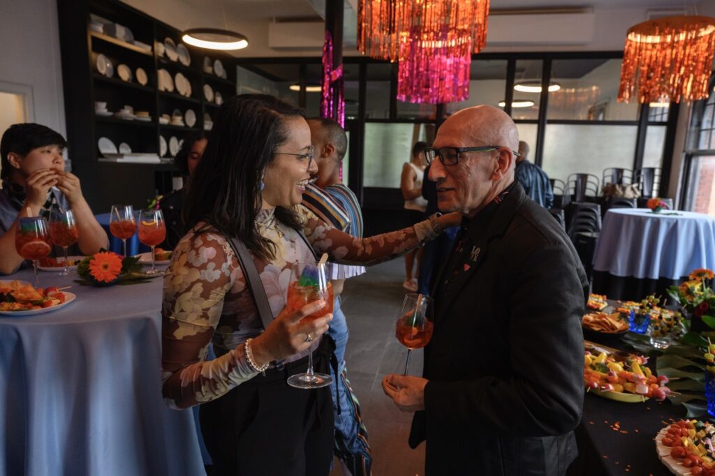 A woman holding a pride popsicle cocktail greets a man in a black long sleeve with glasses.
