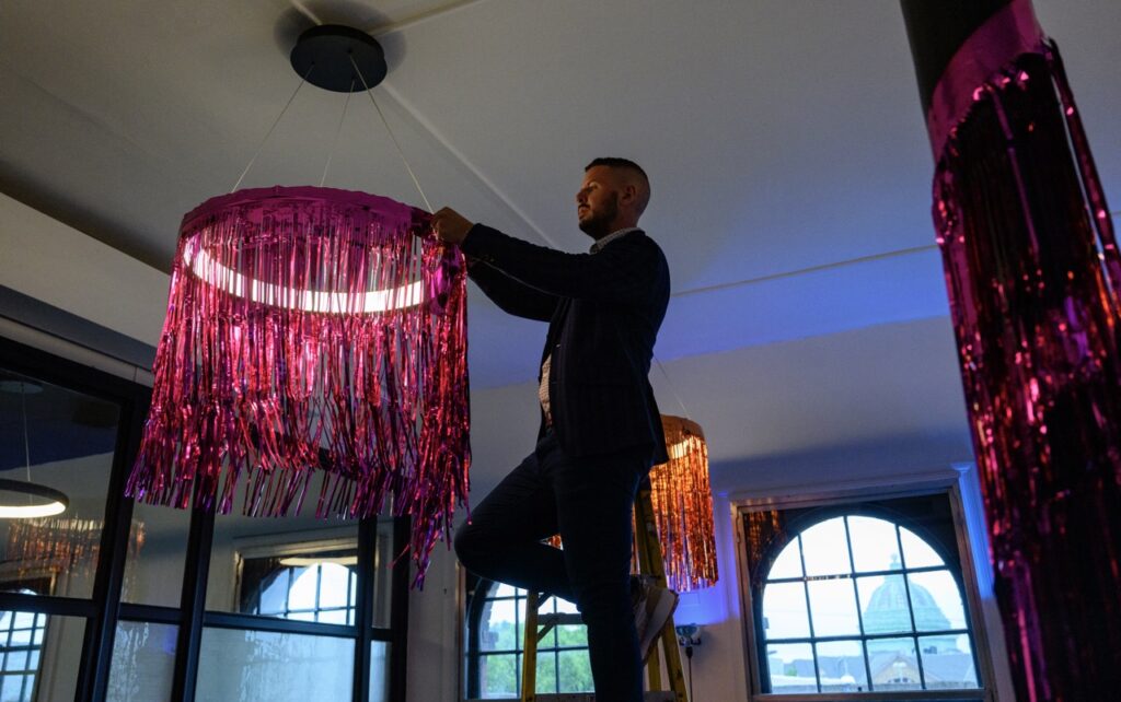 A man in a suit climbs on a ladder to hang up pink decor around a circular light.