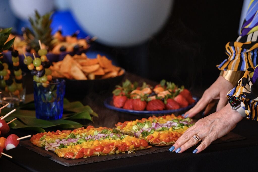 A woman places two rainbow veggie flatbread pizzas on a table with other pride foods.