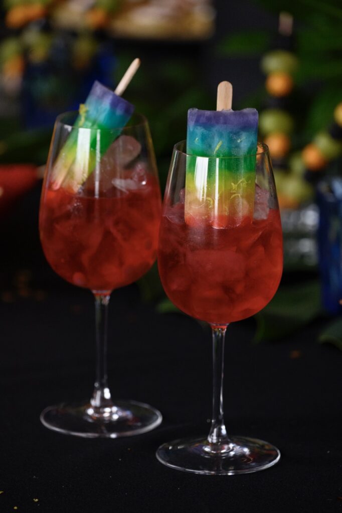 Two wine glasses hold a red cocktail with a rainbow popsicle sticking out of each.