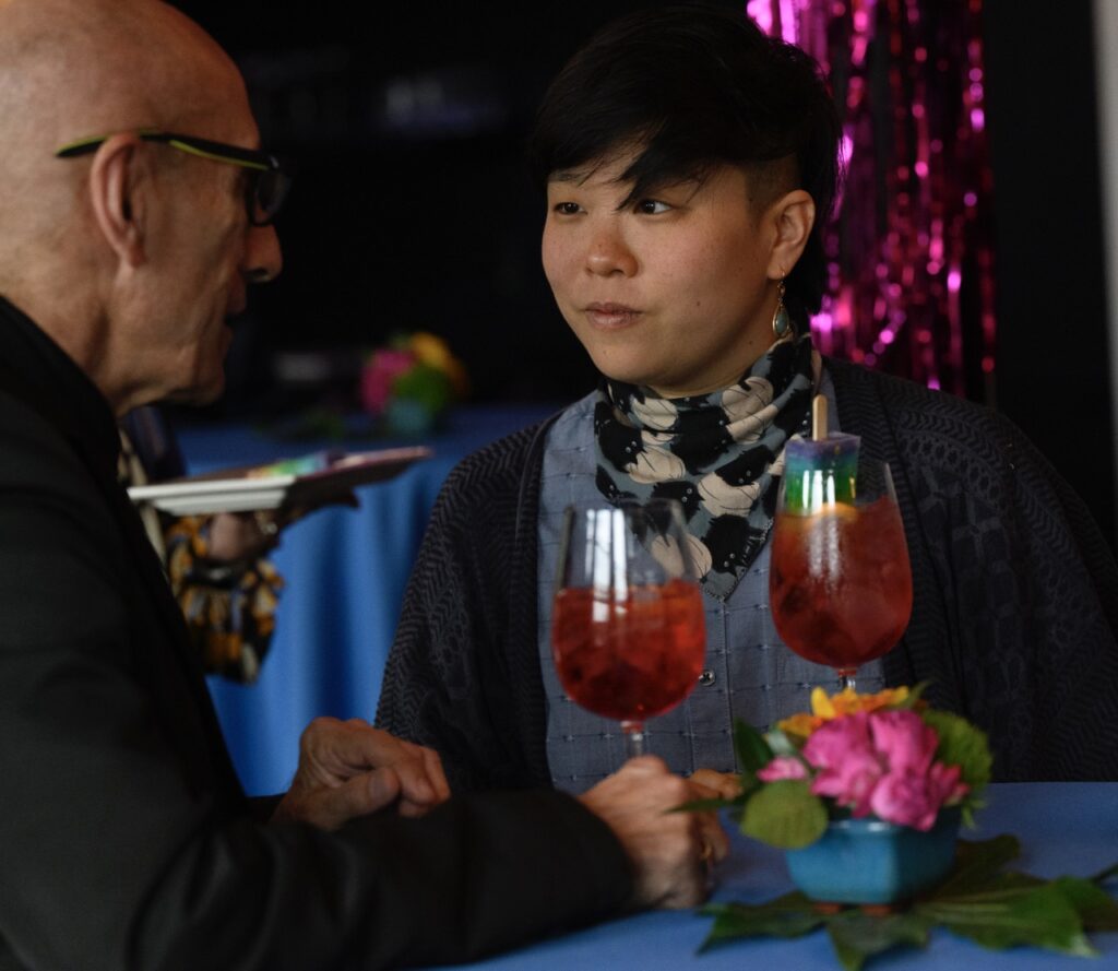 A person in a neck scarf holds a wine glass with a red cocktail and rainbow popsicle while talking to a man in a black shirt.