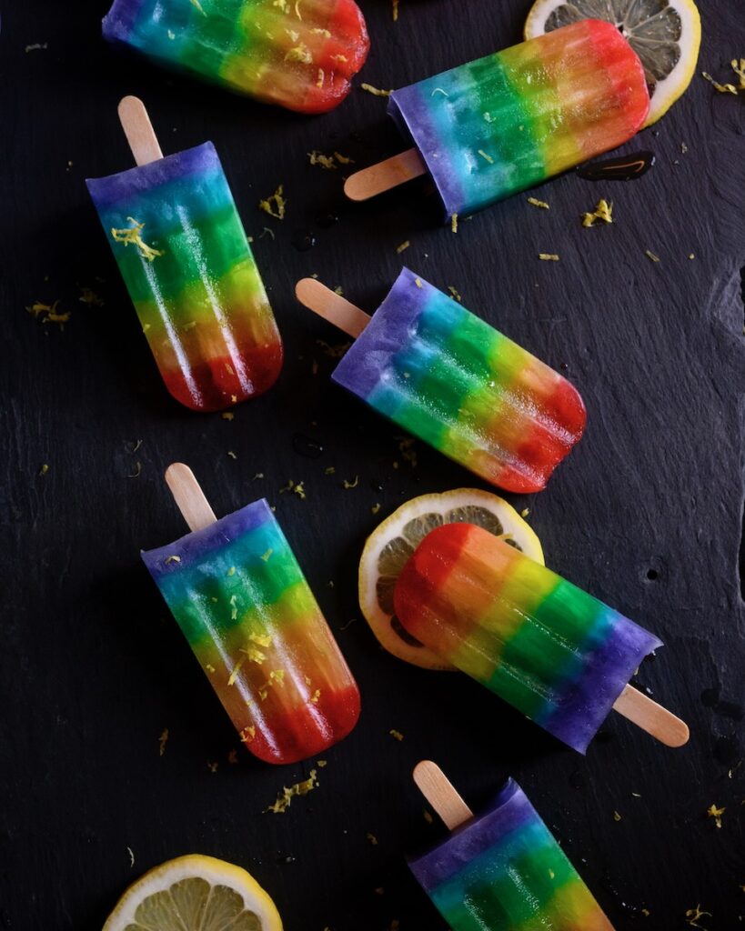 7 rainbow pride popsicles sit on a black table with lemon slices sitting beneath a few of them.