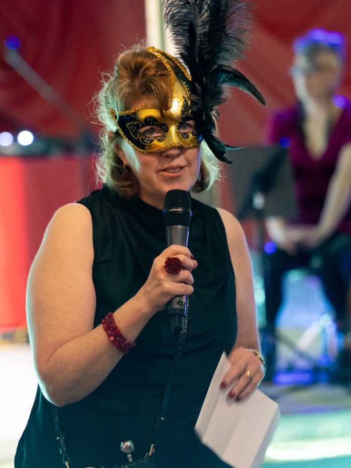 A woman with a gold and black mask speaks into a microphone.