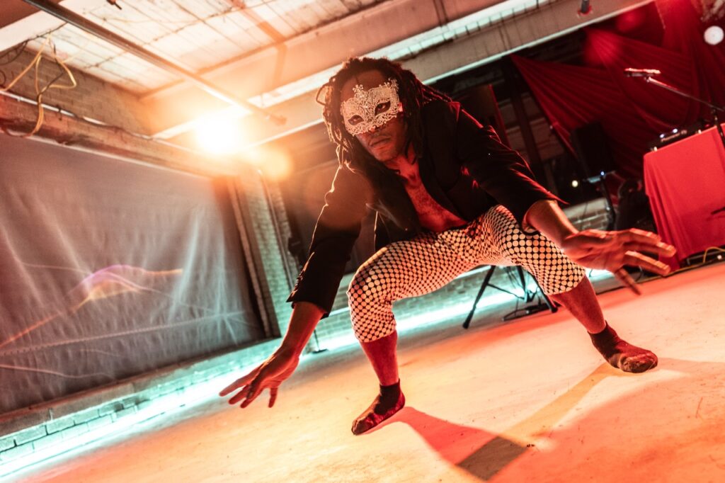 A person squats down in a silver masquerade mask with lights shining in the background.