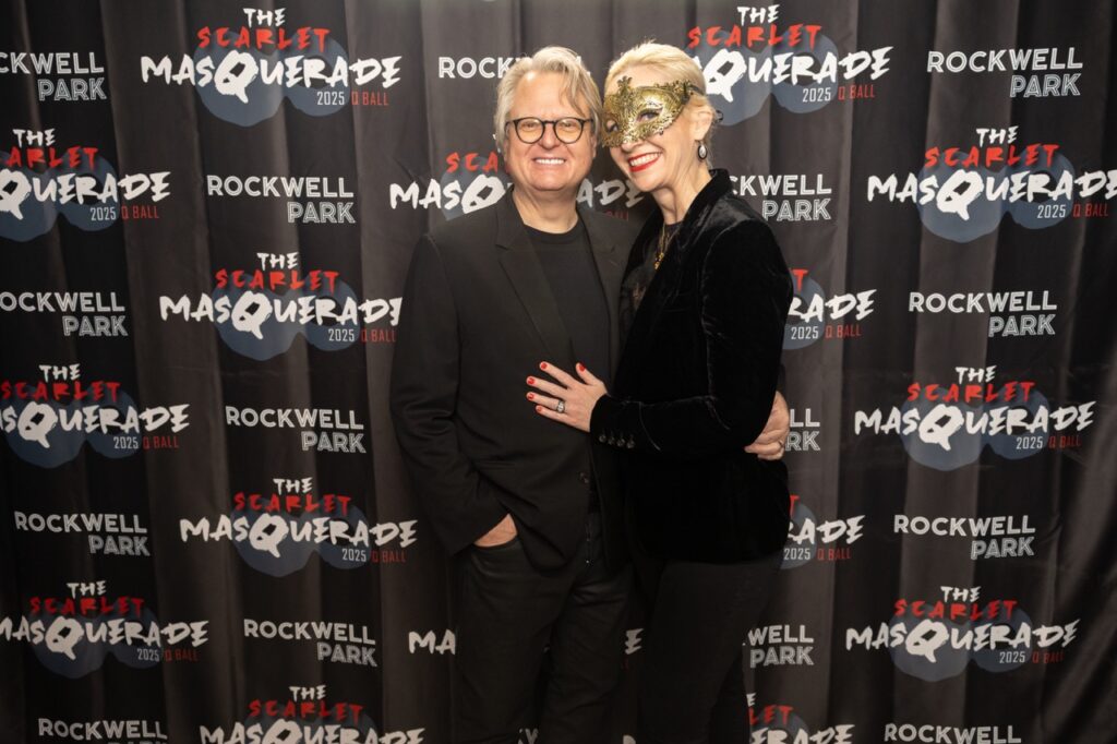 A man in a black suit jacket and woman in a black jacket and gold mask stands together.