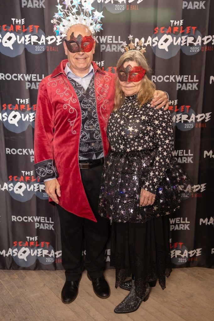 A man and woman in masks pose in front of a photo op background.