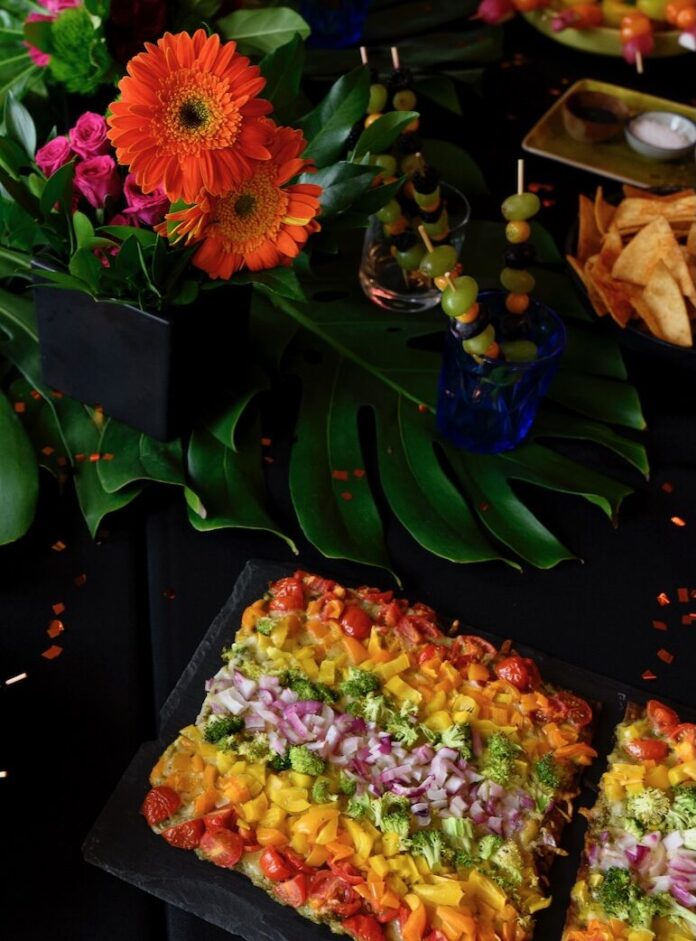 4 Rainbow Recipes for Pride Month A rainbow flatbread pizza sits below an array of bright colorful flowers with fruit skewers sittin in cups to the right side.