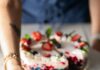 Party Ideas for Your Fourth of July Celebration A red, white, and blue gelatin cake decorated with fresh strawberries and blueberries sits on a platter while a woman holds out a cut slice on a small white plate.