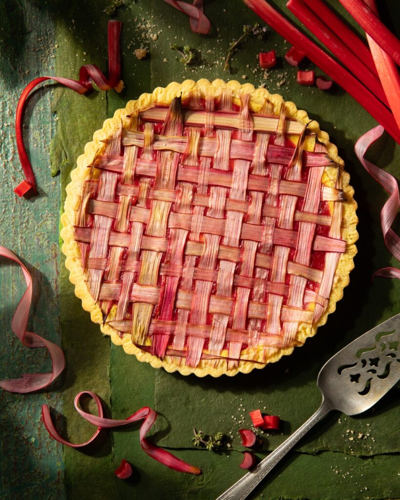 A whole Rhubarb Frangipane Tart with latticed rhubarb slices on top sitting on a green table with a pie spatula nearby.