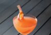 Rhubarb Gimlet A cocktail glass with an orange rhubarb gimlet featuring a rhubarb ribbon garnish all on a black picnic table.