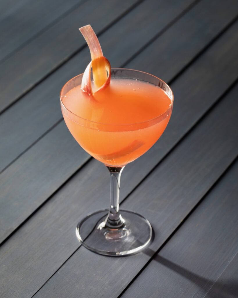 A cocktail glass with an orange rhubarb gimlet featuring a rhubarb ribbon garnish all on a black picnic table.