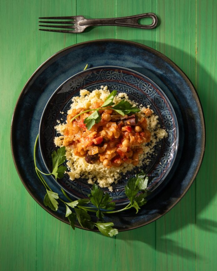 Two blue plates hold a tagine with chickpeas and rhubarb with a garnish of parsley underneath it, all on a green picnic table.