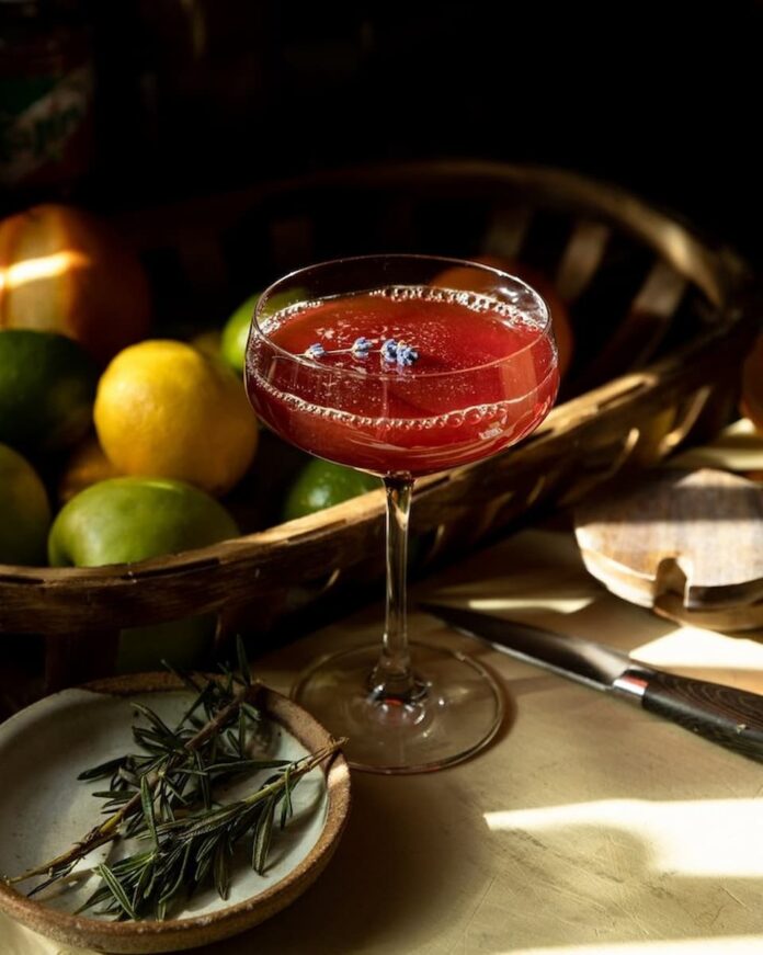 Sangre Sunset Sangre Sunset cocktail sits red in a coupe glass with a toothpick of blueberries floating on top.