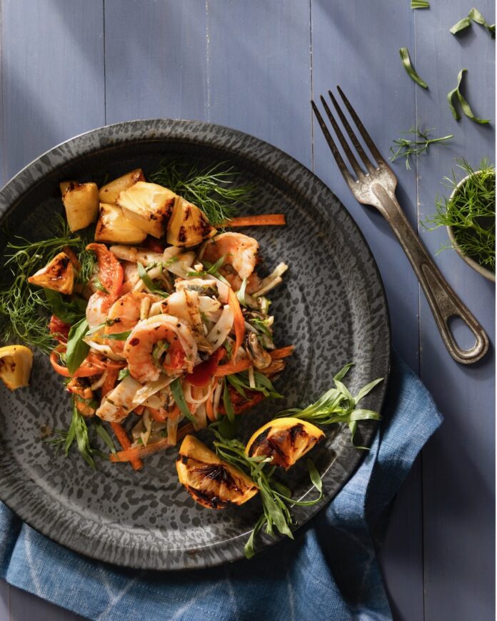 A dark plate holds garden vegetables and seafood in an escabeche salad with a fork and herbs to the right of the plate.