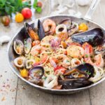 A seafood spaghetti sits in a shallow bowl with shrimp and mussels, all sitting on a wood picnic table.