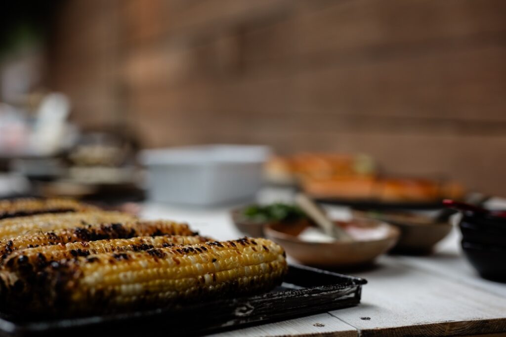 Grilled corn on the cob sits amongst a table of food.