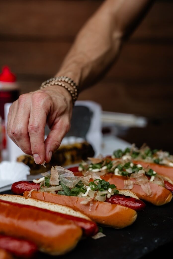 A man sprinkles cheese on top of a gourmet hot dog with greens and other ingredients.