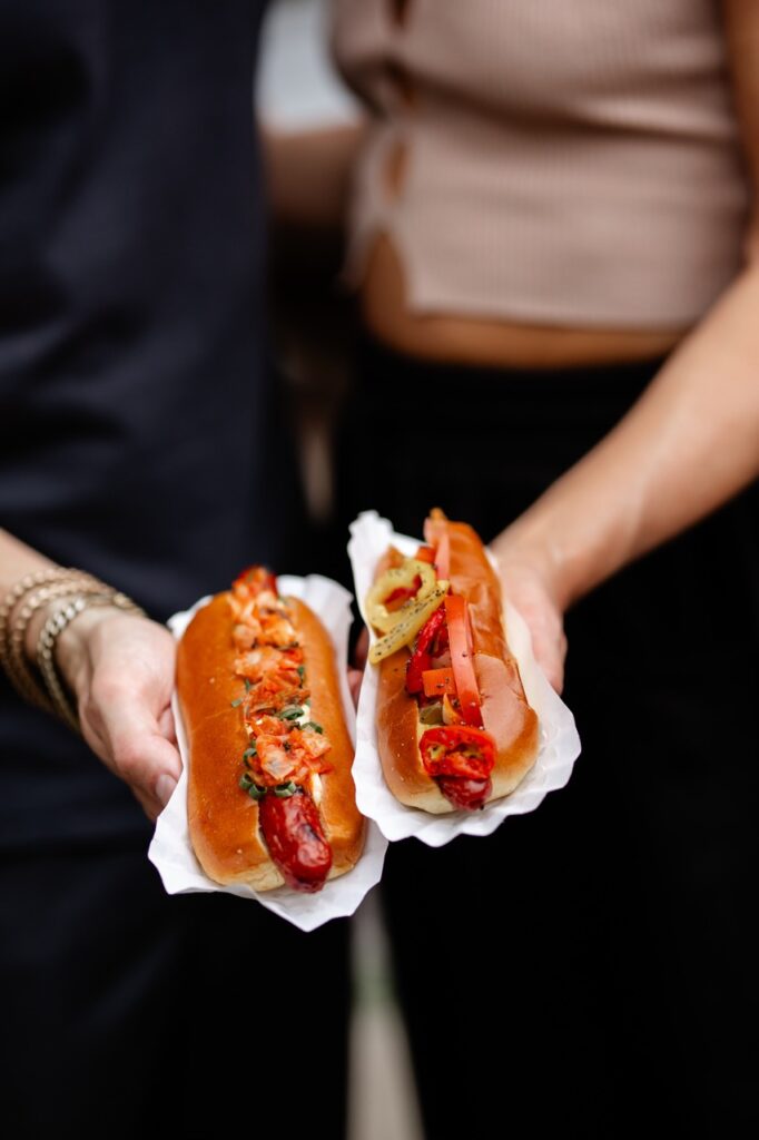 A man and woman each hold a gourmet hot dog in their hand with a piece of paper under the dogs.