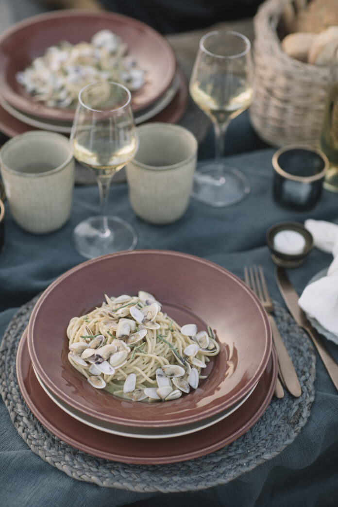 Spaghetti with Clams A red bowl with Spaghetti with Clams sits on a greenish tablecloth with a wine glass, candles, and another bowl of pasta behind it.