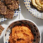 An orange tomato hummus sits in a polka dot and white bowl on a picnic table below another bowl of hummus and a plate of wheat crisps.