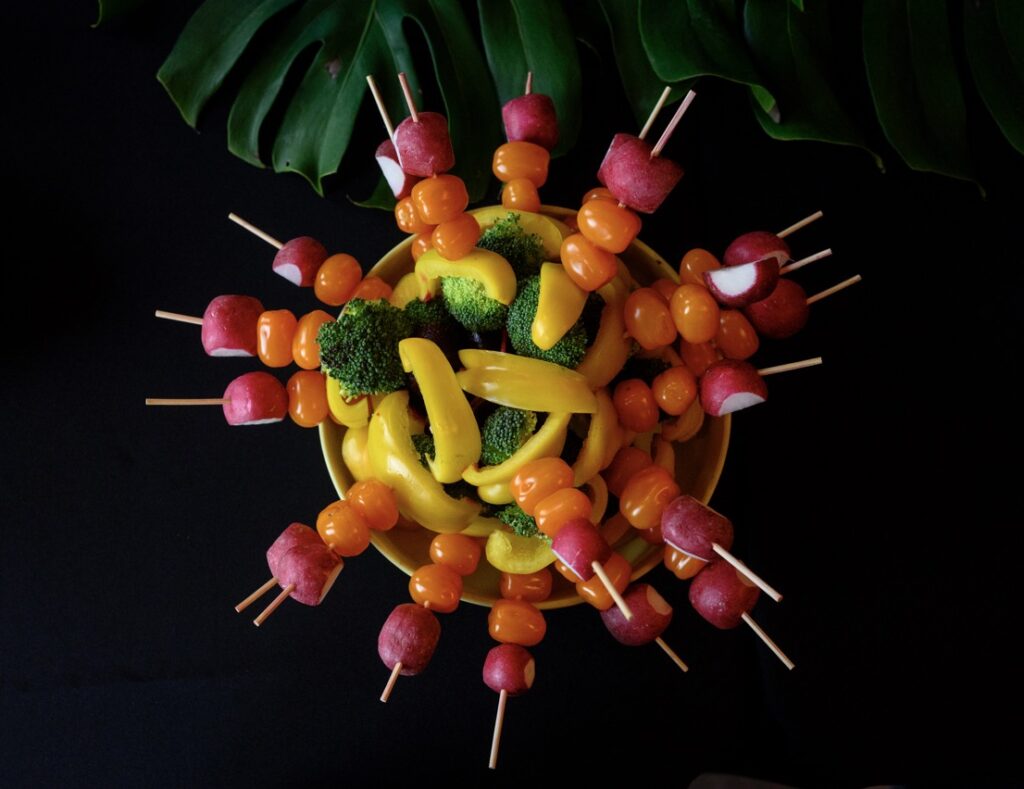 Various rainbow vegetable skewers sit in a large bowl on a black table.