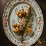 A whole roasted branzini with fingering roasted potatoes sits on a white decorated plate with bits of orange and green flowers.