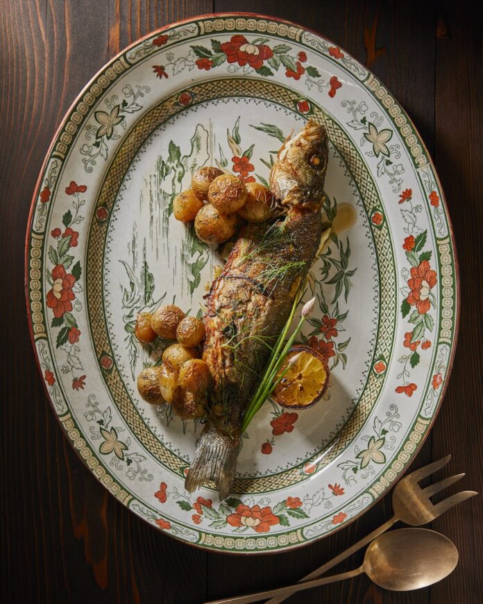 Whole Roasted Branzini A whole roasted branzini with fingering roasted potatoes sits on a white decorated plate with bits of orange and green flowers.