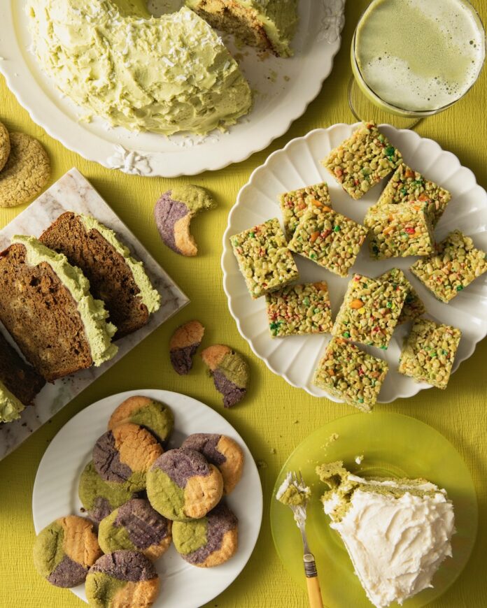 Three white plates and various other platters hold matcha dessert recipes like tricolor cookies, banana bread, chiffon cake, rice krispies treats, cake, and a martini, all on a green table.