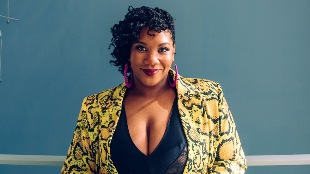 A woman in a yellow print jacket stands in front of a blue background.