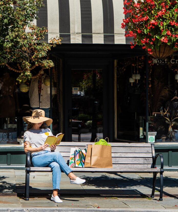 A woman sits on a bench outside an Appalachia city shop reading as her bags sit beside her.